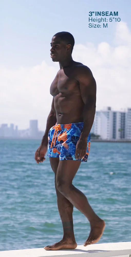 Man wearing colorful swim shorts standing on a dock with water and buildings in the background