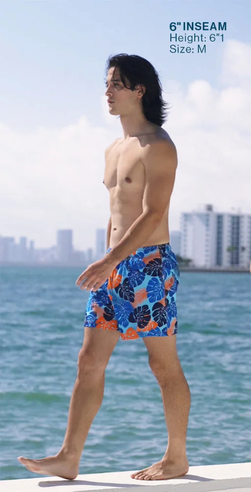Man wearing colorful swim shorts standing by a waterfront with a city skyline in the background.