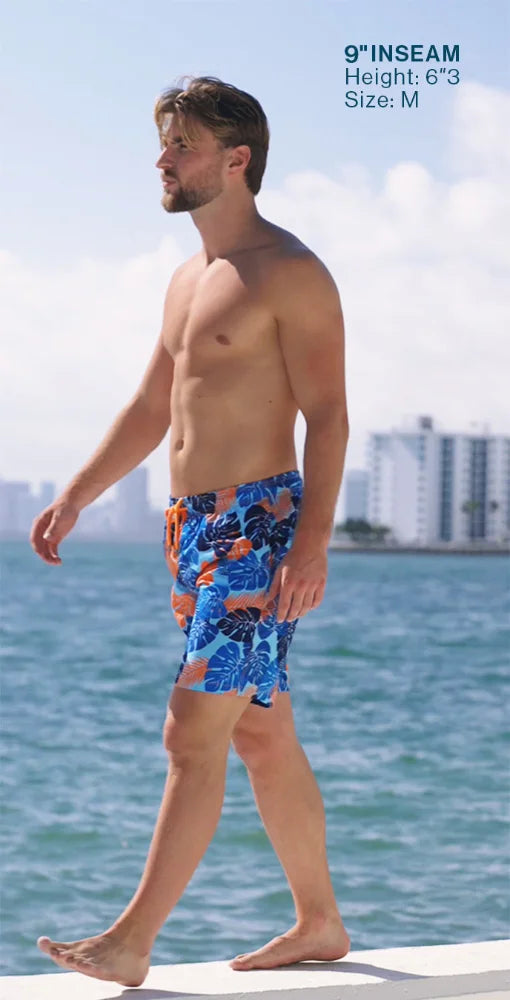 Man wearing colorful swim shorts standing on a dock with water and buildings in the background