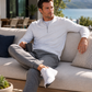 A man sits on a light beige outdoor sofa wearing a white long-sleeve quarter-zip pullover, gray jogger pants, and white sneakers, holding a black mug with a scenic lake view in the background.