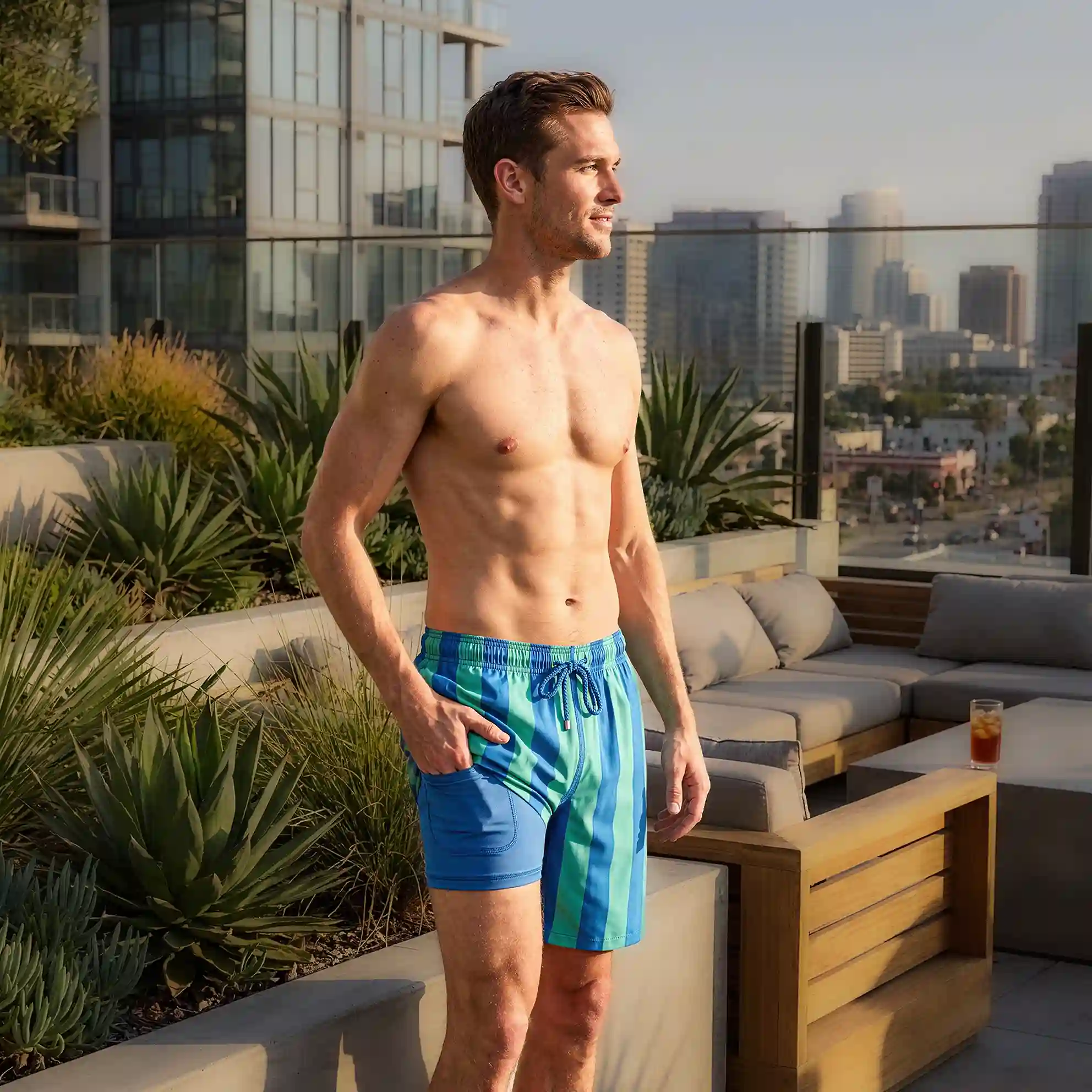 Man on a rooftop terrace above the city skyline in Coastal Cabana Ocean and Palm hybrid swim shorts, blue and green cabana stripe