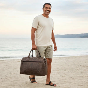 Man holding a brown leather bag on a beach with ocean and sky in the background