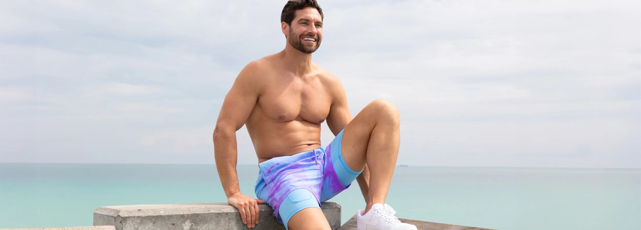 guy on a ledge with a background of the ocean while wearing white shoes and color changing swim trunks