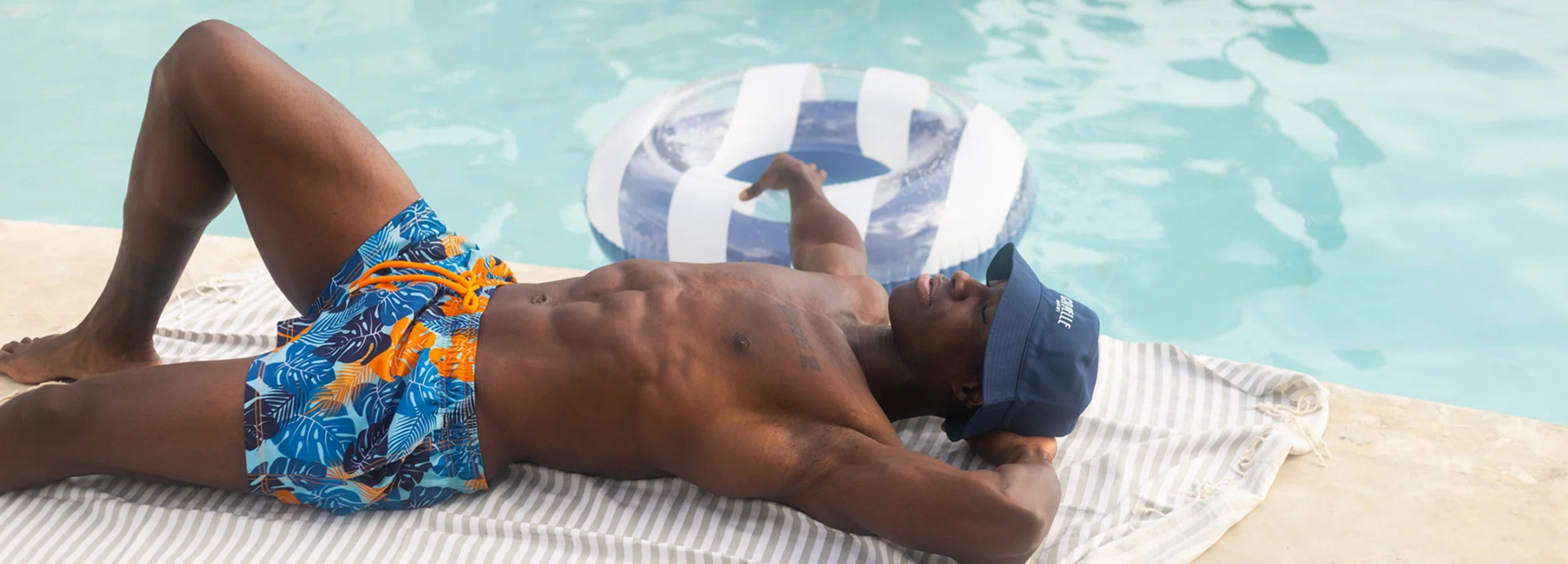 guy lounging on his back with a beach towel next to the pool while wearing a capelle bucket hat and blue jungle swim trunks