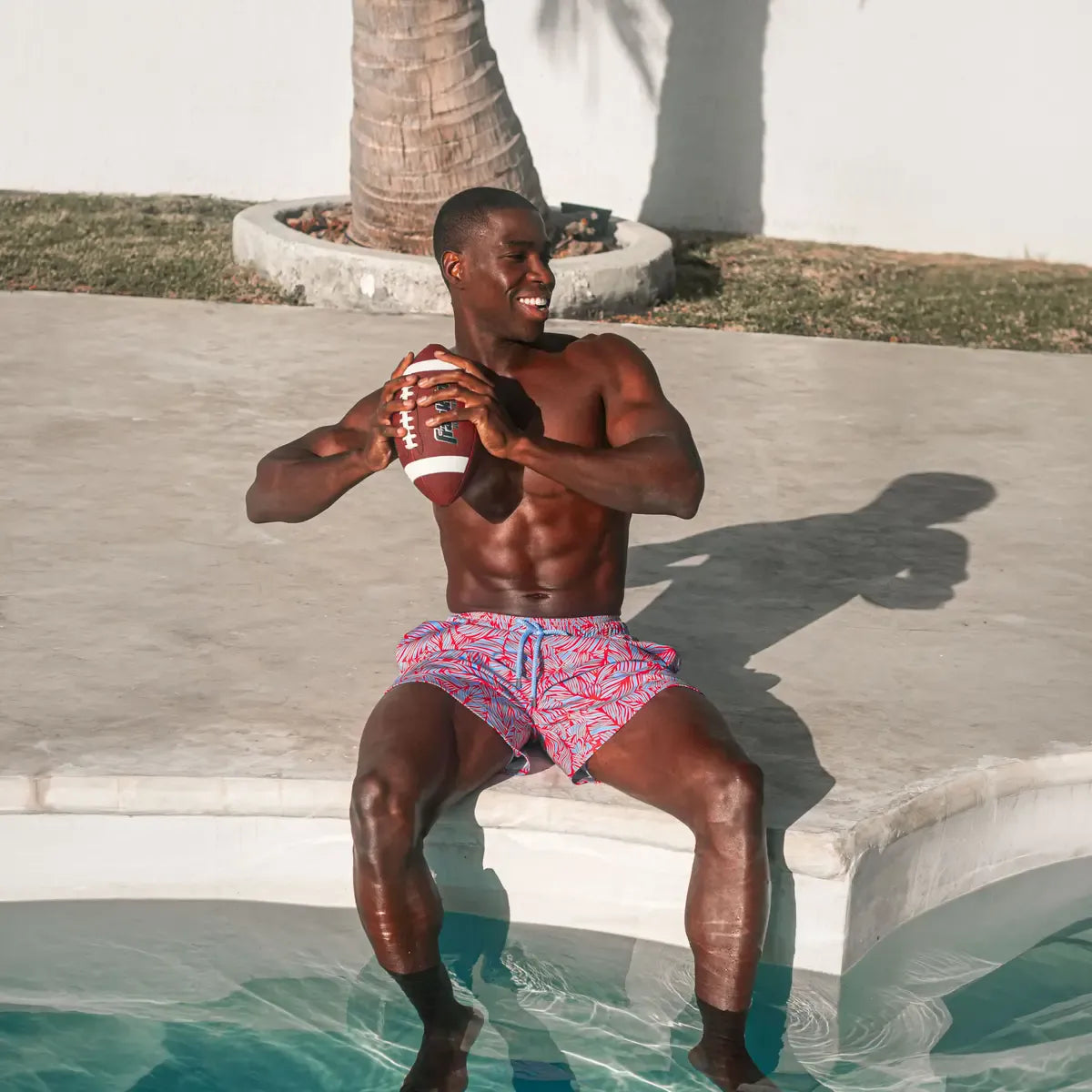 Man holding a football at the edge of a pool in Captive hybrid swim shorts, a coral and pale blue tropical leaf print