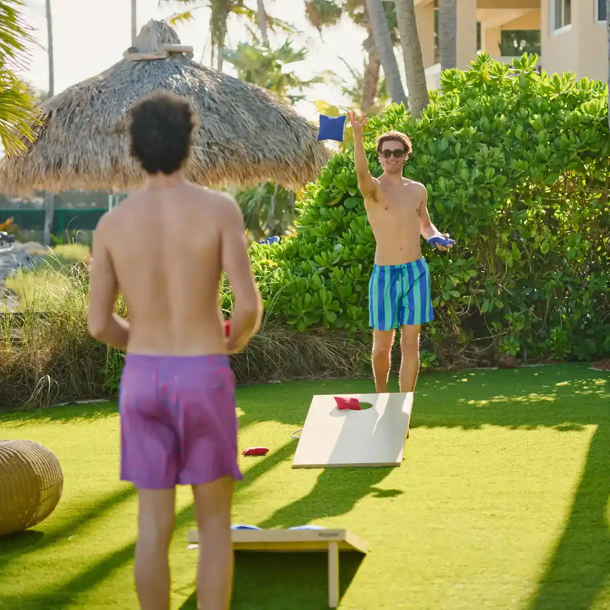 Man in Coastal Cabana Ocean and Palm hybrid swim shorts, a blue and green cabana stripe, playing cornhole on a lawn under a thatched umbrella