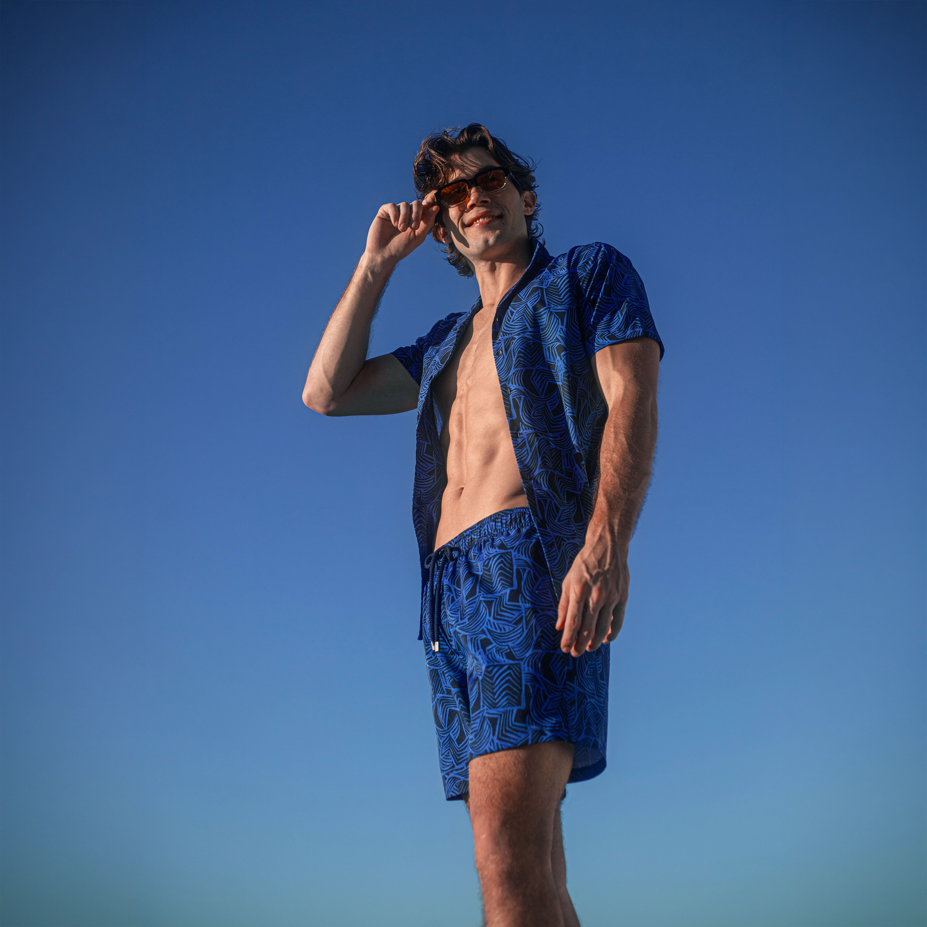 Man against a clear blue sky in an Enigma resort shirt and matching royal blue leaf print hybrid swim shorts