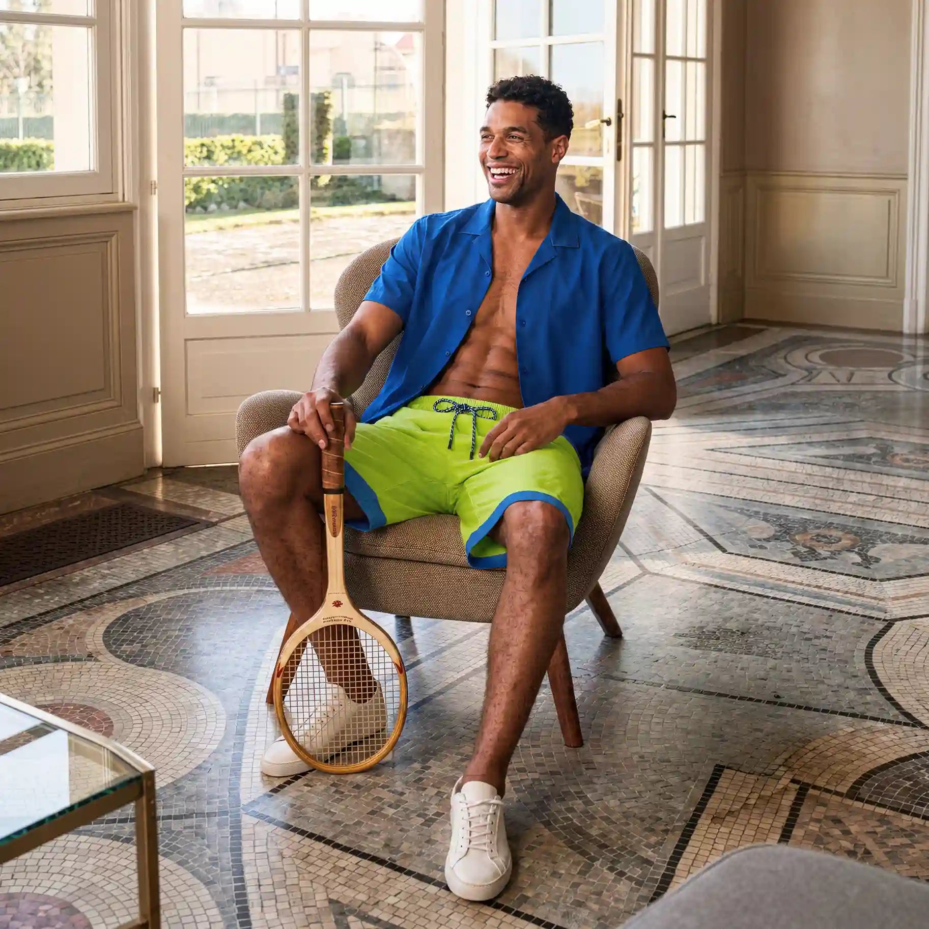 Man seated in an armchair by a sunlit window in Key Lime hybrid swim shorts and a blue resort shirt