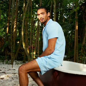 Man seated among bamboo in a Blue Azure resort shirt and matching pale blue hybrid swim shorts