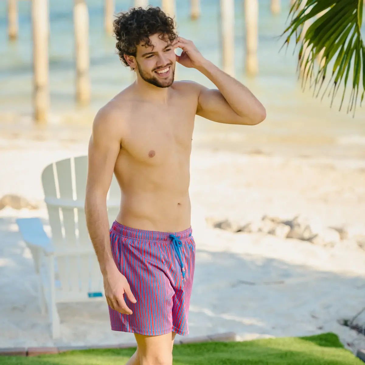 Man walking on a lawn beside the beach in Linear Drift Blaze and Harbor hybrid swim shorts, coral red with a blue wave stripe