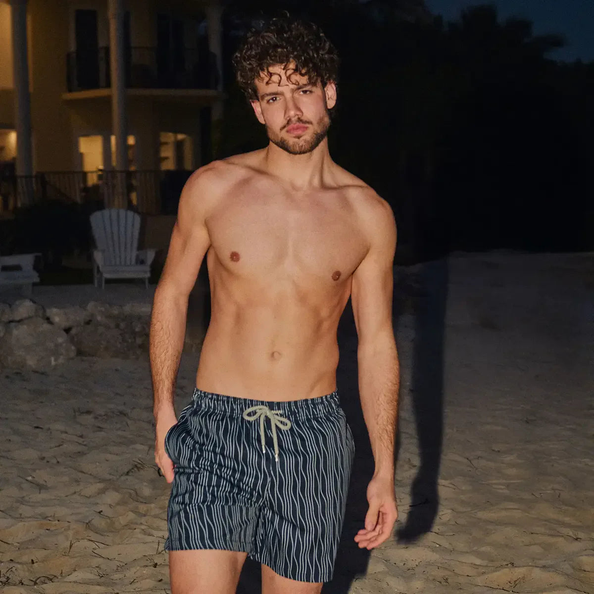 Man in Linear Drift Storm and Fog hybrid swim shorts, charcoal with a white wave stripe, on the sand at dusk