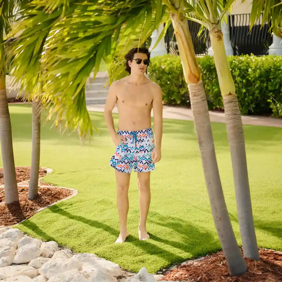 Man in Line Rush Ocean and Sol hybrid swim shorts, a navy, aqua and coral zigzag print, barefoot on a resort lawn under palm trees