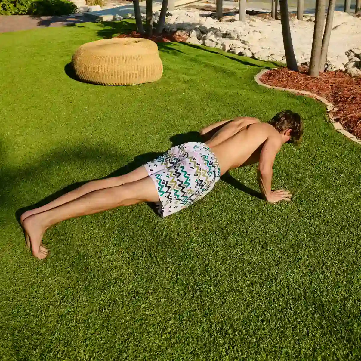 Man holding a plank on a lawn in Line Rush Orchid and Kelp hybrid swim shorts, a green, olive and pink zigzag print