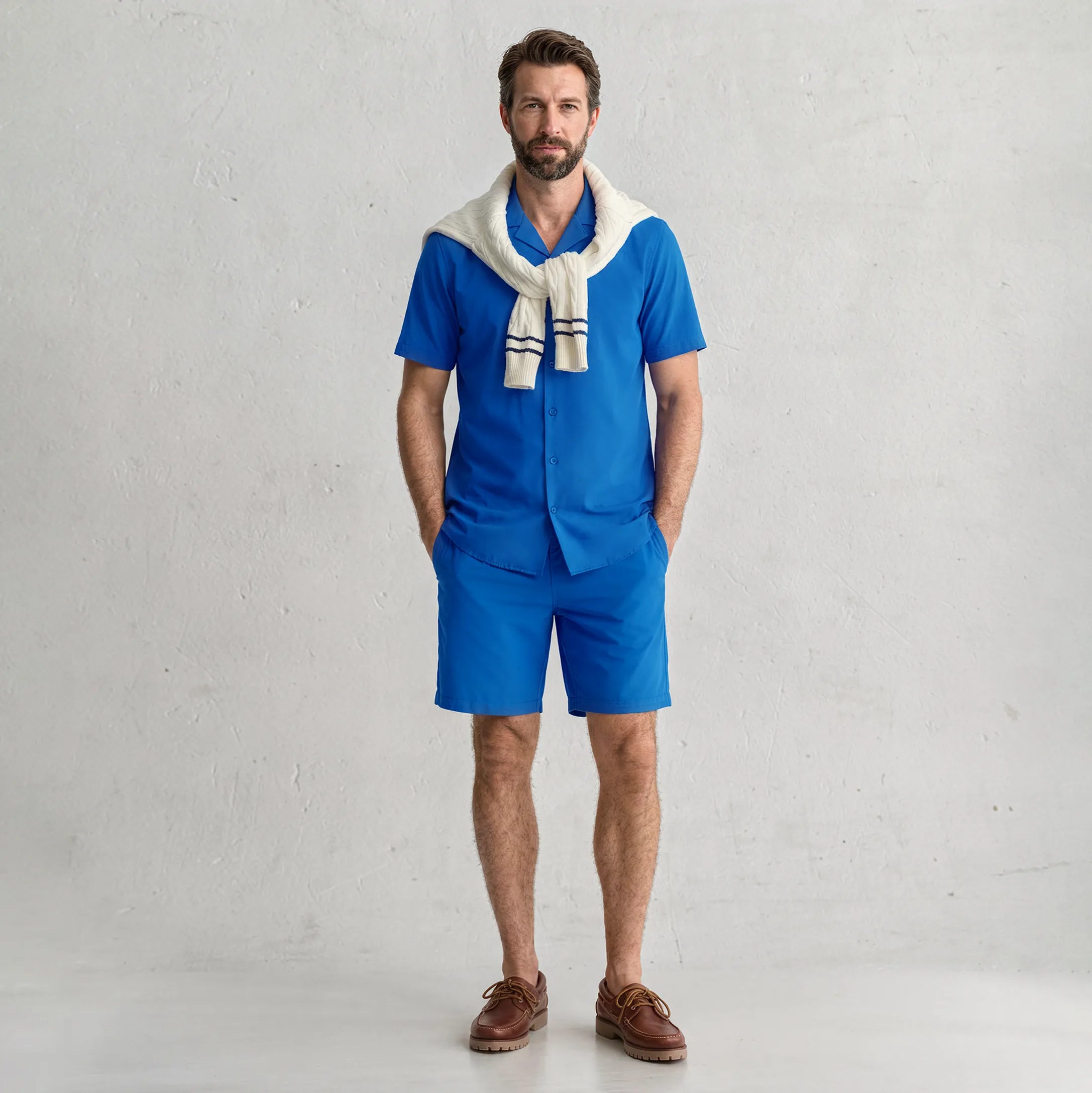 Man in a matching Ocean Blue resort shirt and hybrid swim short set with a cream sweater over the shoulders, studio view