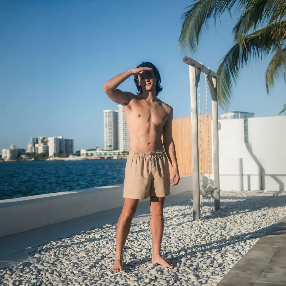 Man in Sahara Dunes linen resort shorts in sand beige, on a pebbled waterfront terrace with a city skyline across the water