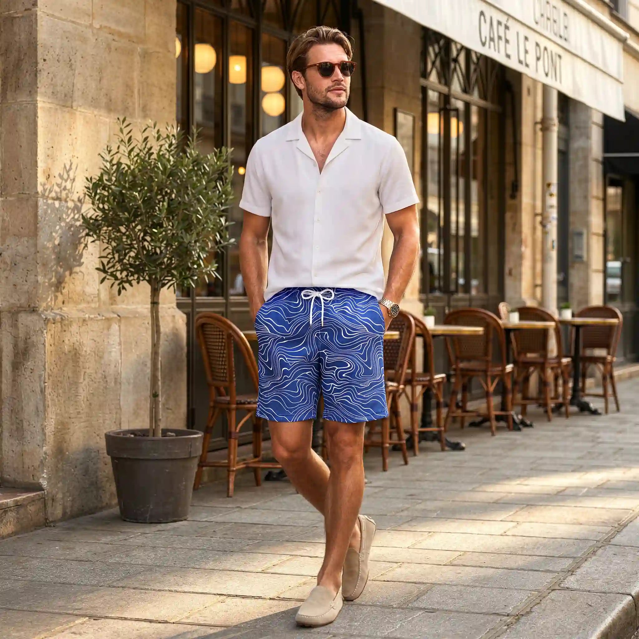 Man walking past a European cafe in Sand Waves Harbor and Surf hybrid swim shorts and a white Arctic White resort shirt