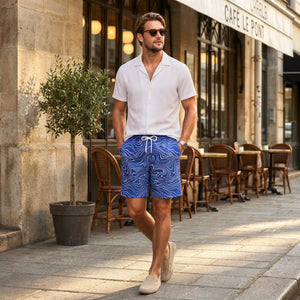 Man walking past a European cafe in Sand Waves Harbor and Surf hybrid swim shorts and a white Arctic White resort shirt