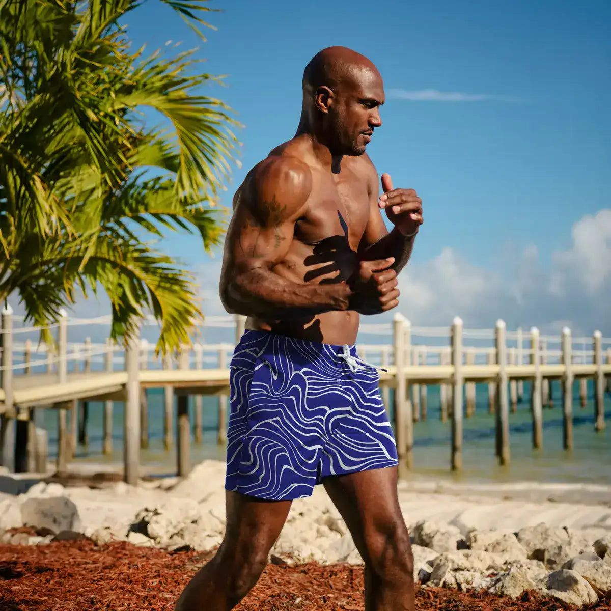 Man in Sand Waves Harbor and Surf hybrid swim shorts, cobalt with a white wave line print, on the sand beside a wooden pier