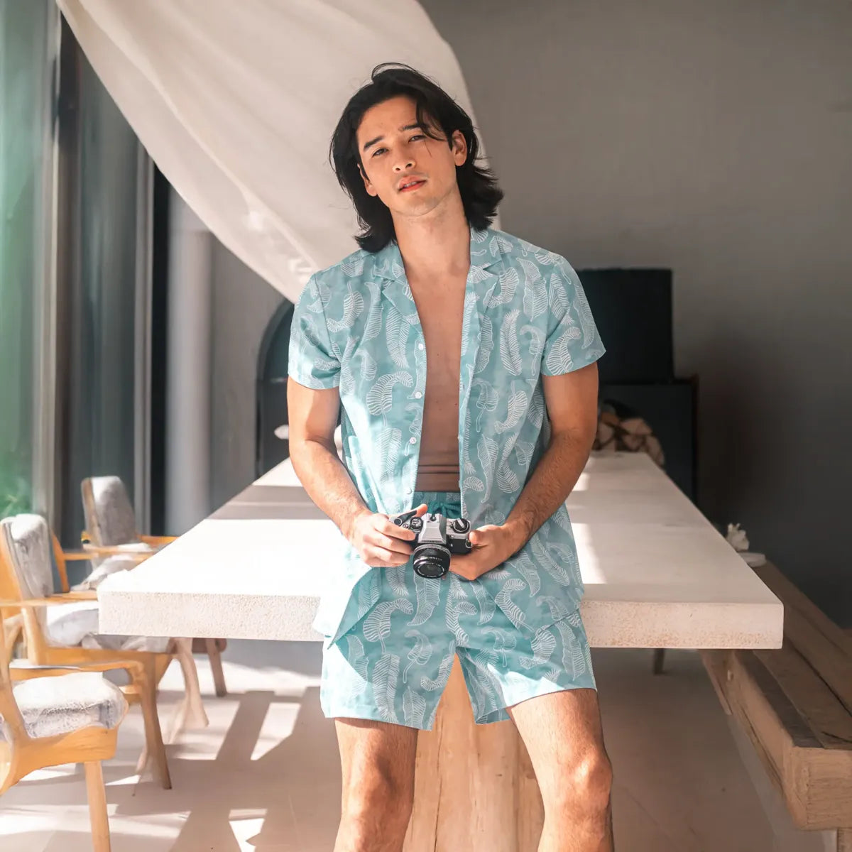 Man indoors by linen drapes in a Serenity resort shirt and matching pale aqua hybrid swim shorts