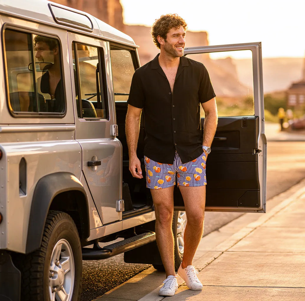 Man stepping out of a vintage Land Rover in Sunset Spritz Surf and Storm hybrid swim shorts and a black resort shirt