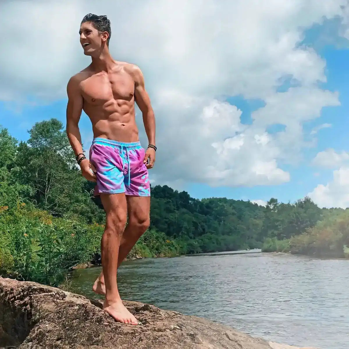 Man standing on a rock above a river in Switch Blue/Purple color changing hybrid swim shorts, purple turning blue where wet